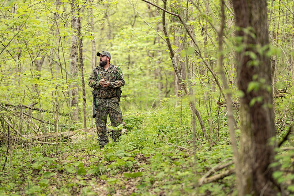 Man in camo using a turkey call in green wooded land.