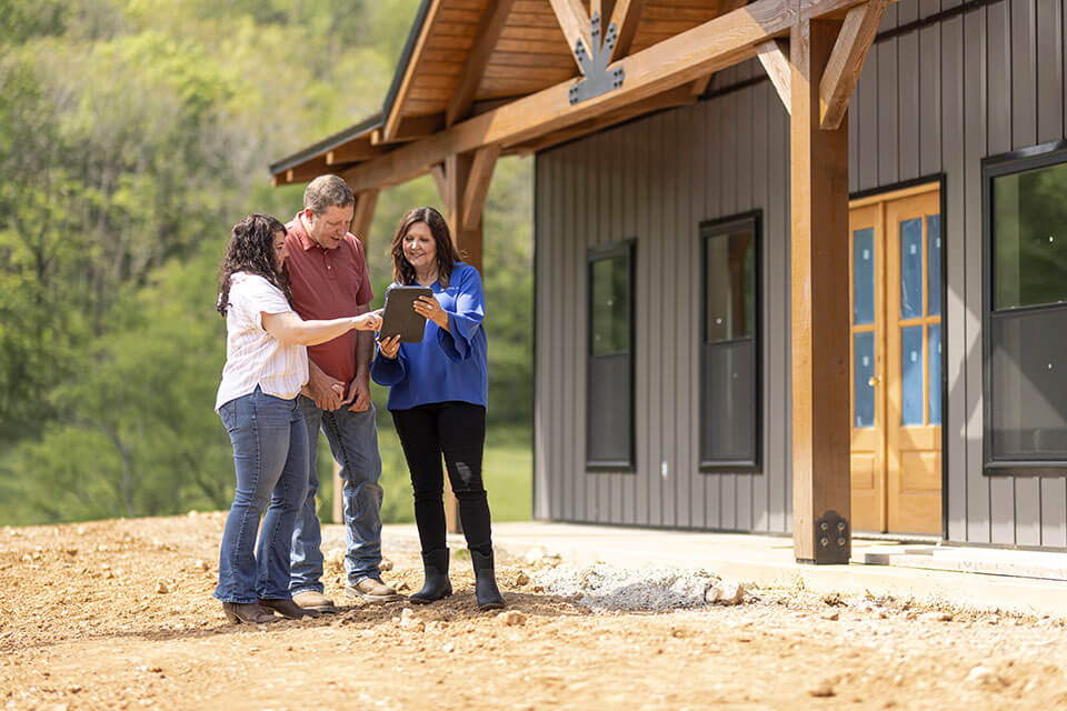 A woman shows something on a tablet screen to a man and woman outside a newly constructed barndominium.