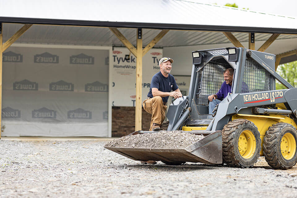 A man in a skid steer loader laughs with a man on a building site.