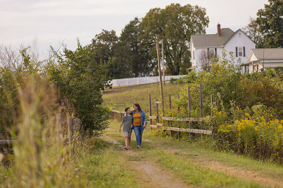 A woman and child walk arm in arm down a grassy gravel path with a house in the distance.