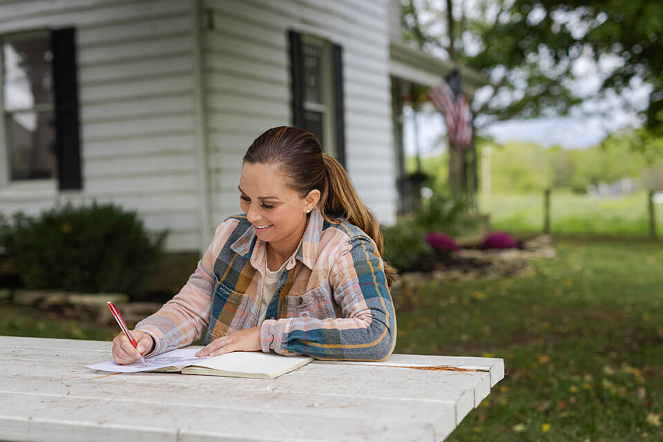 A woman writes in a notebook on a picnic bench outside a home.